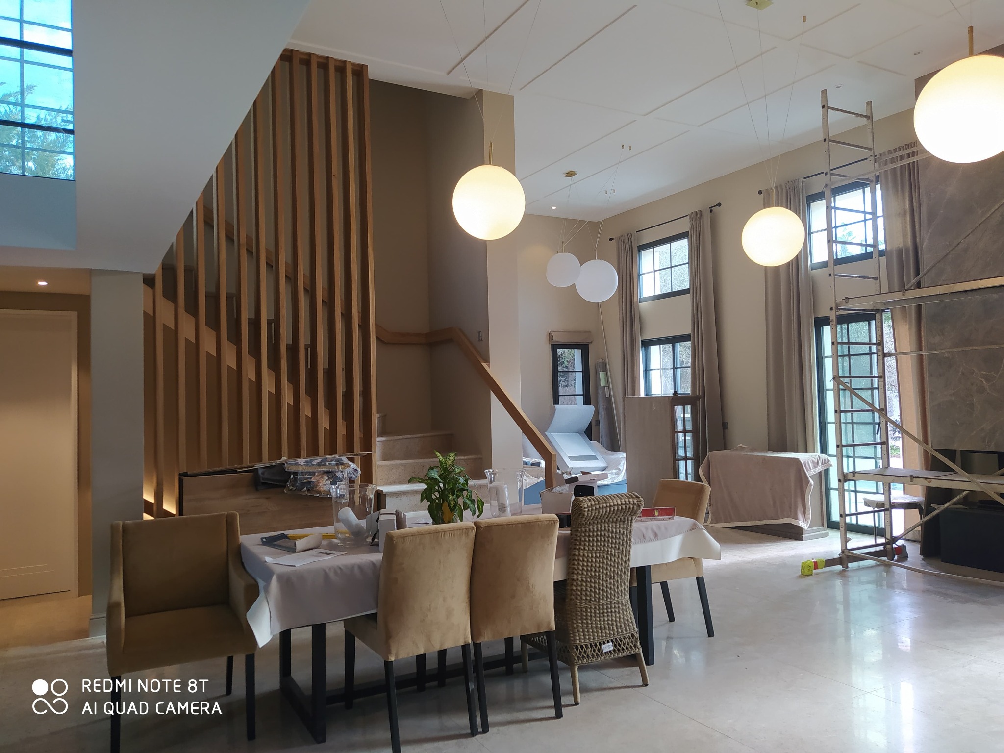 Jasne wnętrze domu w trakcie wykańczania: drewniane schody z ażurową balustradą, marmurowe podłogi, wiszące lampy kule i rusztowanie przy oknie. Widok na jadalnię.