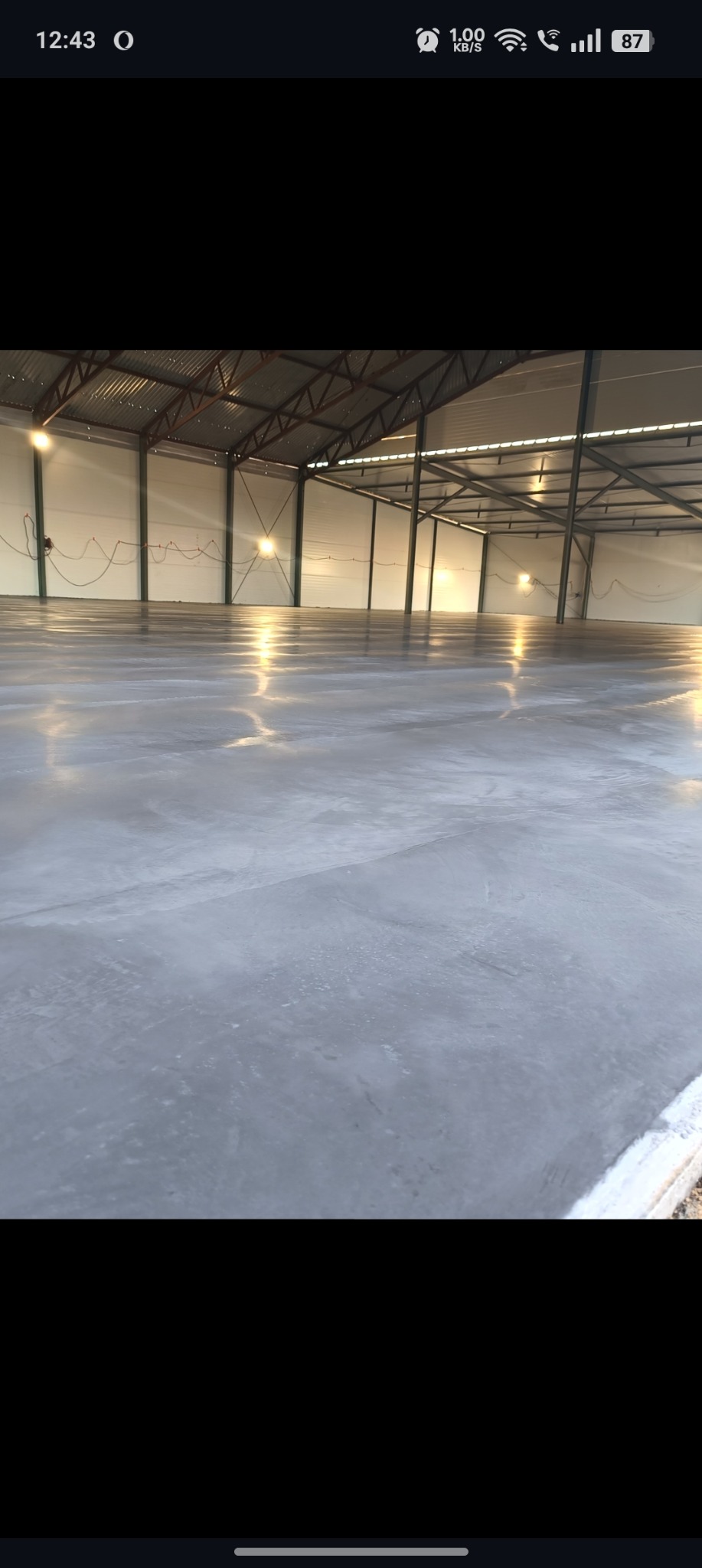 Wnętrze hali z gładką, szarą posadzką przemysłową. Widoczne metalowe elementy konstrukcyjne dachu i oświetlenie. Świeżo wylana, błyszcząca powierzchnia betonu.