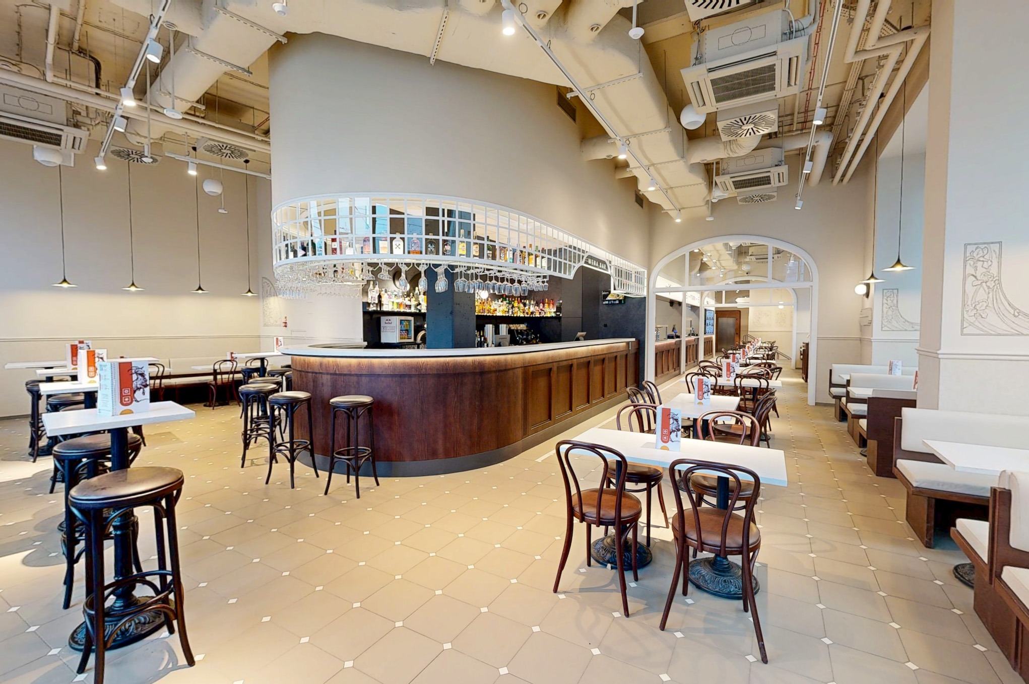 Jasne wnętrze restauracji z drewnianym barem, białymi stolikami i krzesłami. Widoczne oświetlenie sufitowe i dekoracje ścienne w stylu art deco.