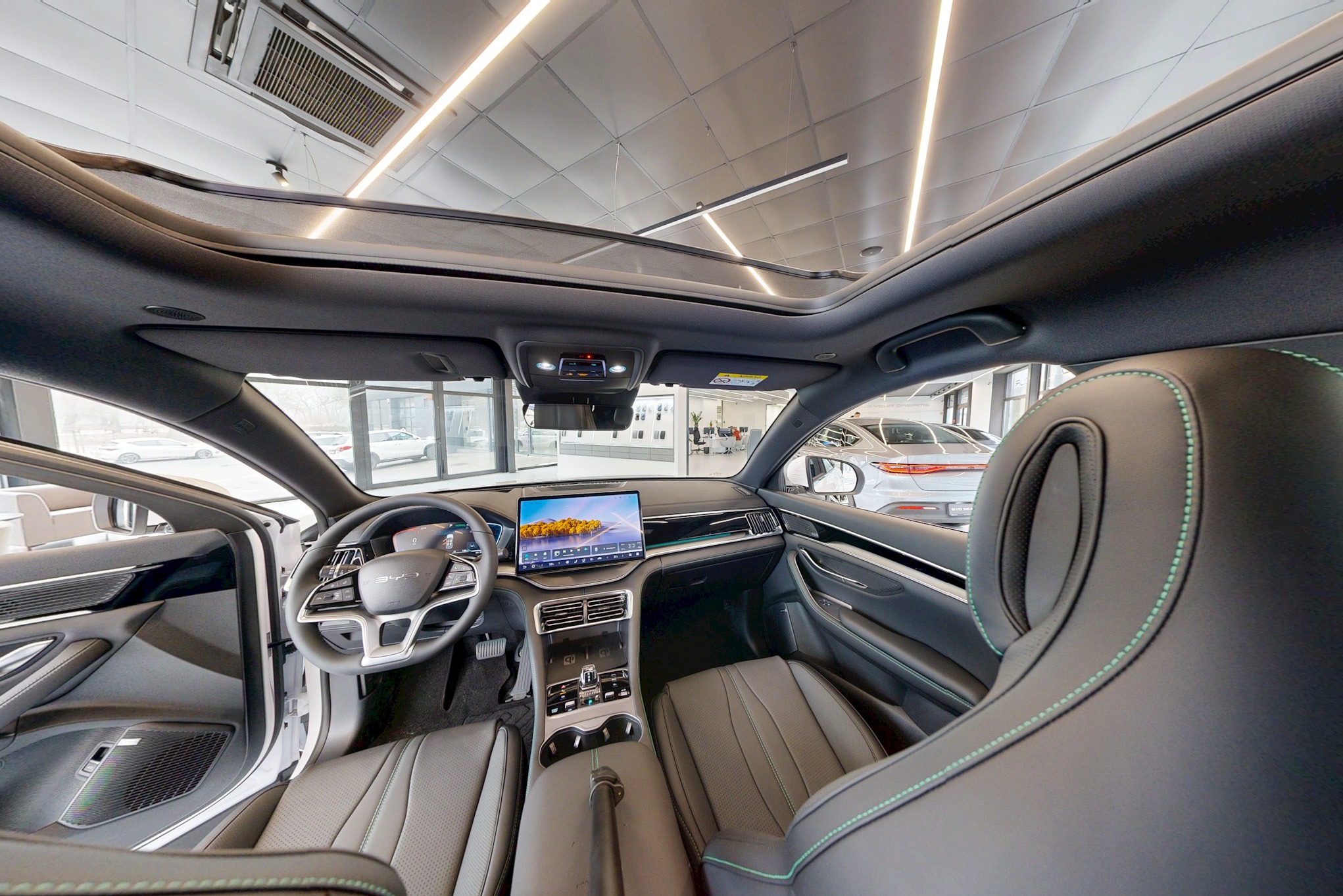 Wnętrze nowoczesnego samochodu z panoramicznym dachem, widok z tylnego siedzenia na deskę rozdzielczą i ekran dotykowy. Skórzana tapicerka z zielonymi przeszyciami.