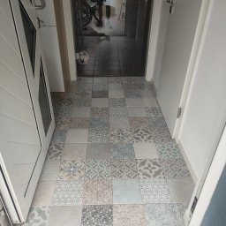 Firma budowlano-usługowa Bud-Mal Tomasz Malczyk - Wzorzysta, patchworkowa podłoga z płytek w przedpokoju, widok z góry. Otwarta biała rama drzwi po lewej, w tle widoczny fragment roweru i piłki.