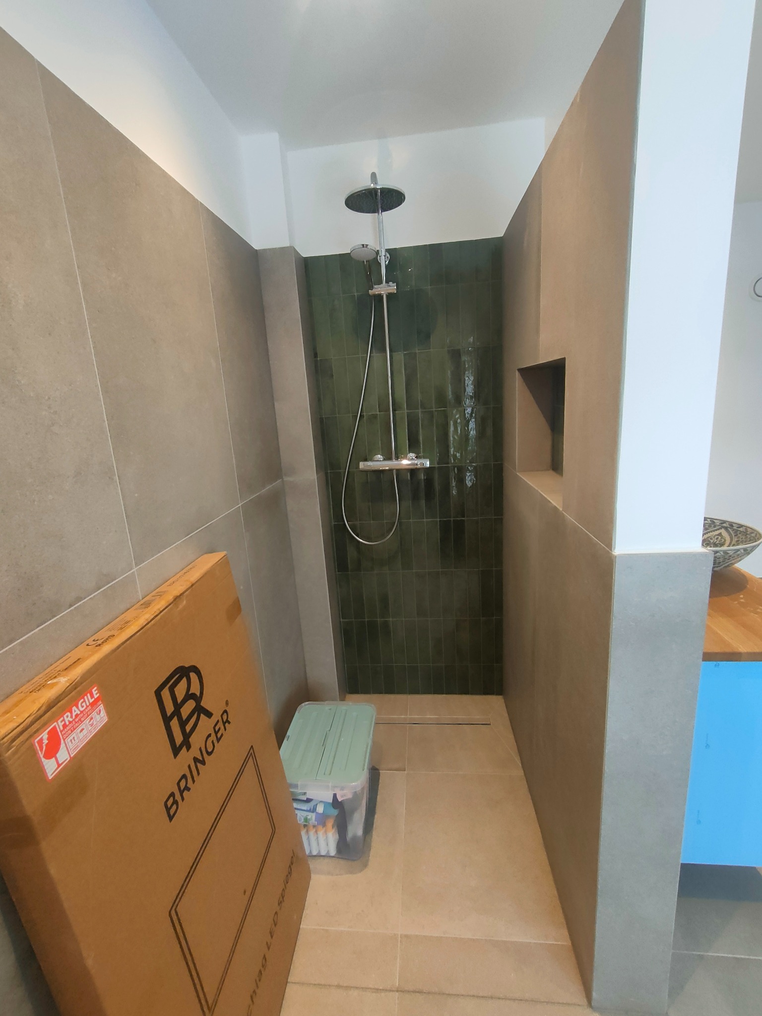 Wykończona kabina prysznicowa z zielonymi płytkami, szarą zabudową i odpływem liniowym. Na pierwszym planie karton z napisem BRINGER i pojemnik z akcesoriami.