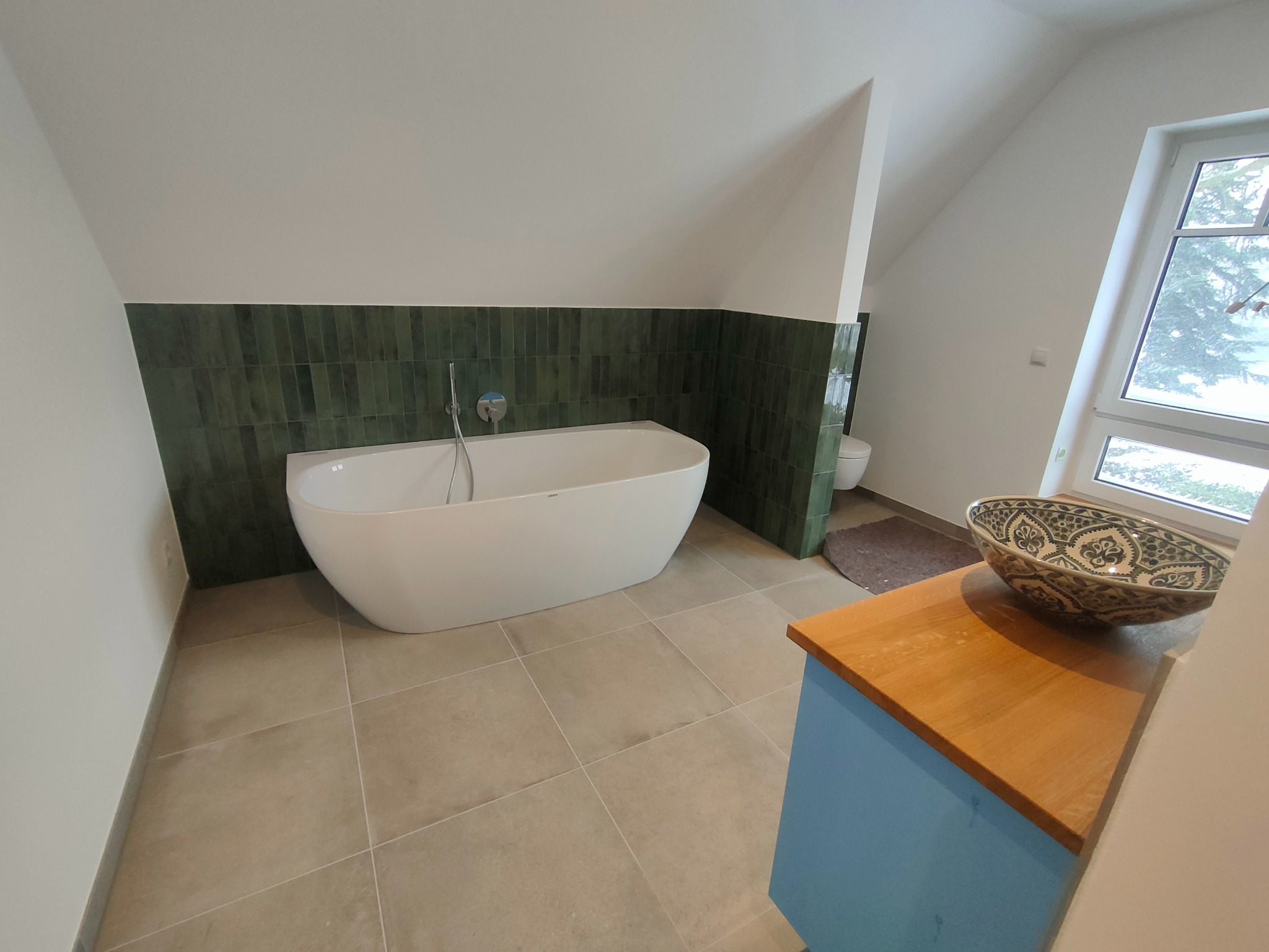 Nowoczesna łazienka na poddaszu z wolnostojącą wanną, zielonymi płytkami ściennymi i szafką z umywalką w stylu marokańskim. Jasne płytki podłogowe i okno.