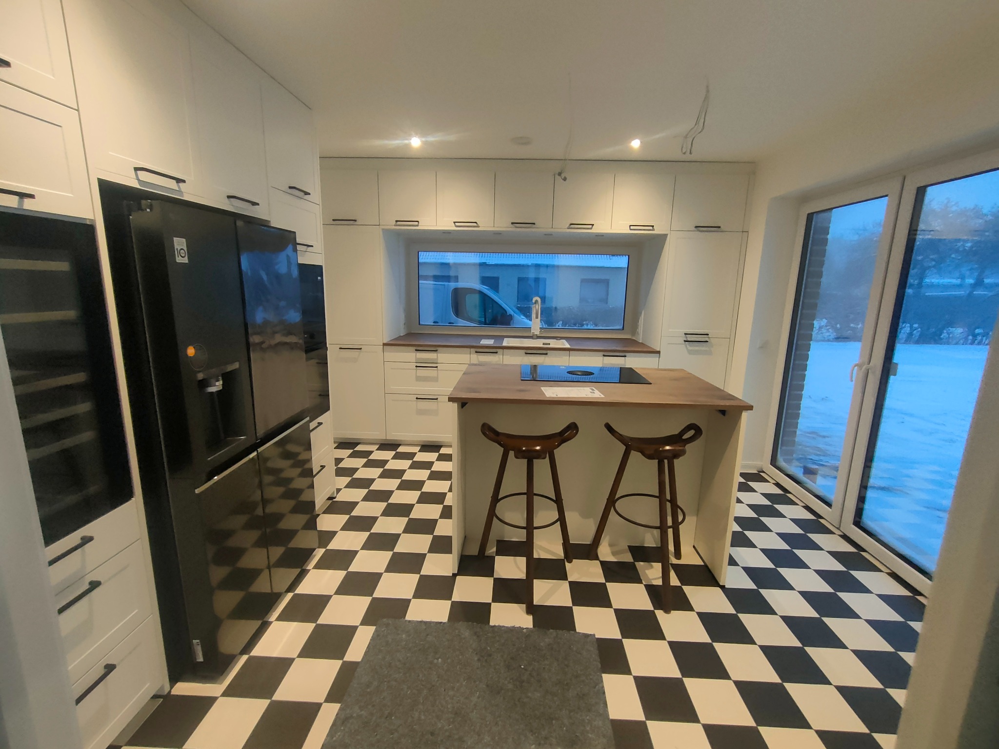 Nowoczesna kuchnia z czarno-białą podłogą w szachownicę, wyspą z drewnianym blatem i białymi szafkami. Duże okno nad blatem roboczym. Minimalistyczny design.