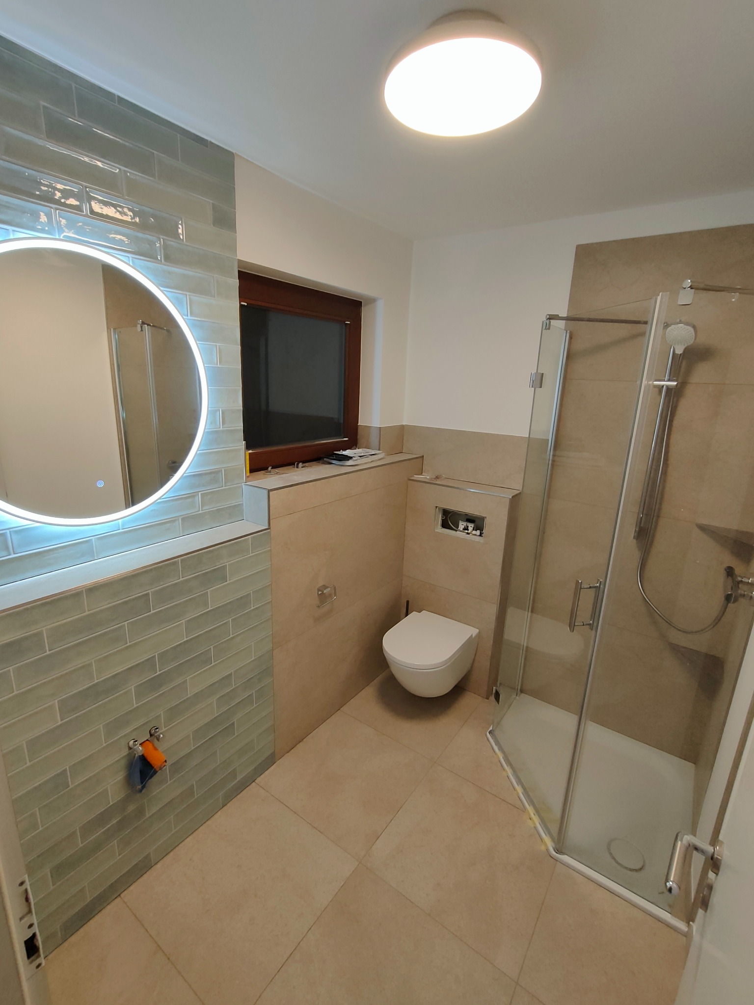 Nowoczesna łazienka z beżowymi i błękitnymi płytkami, okrągłym lustrem z podświetleniem LED i kabiną prysznicową ze szklanymi drzwiami. Widok na toaletę pod oknem.