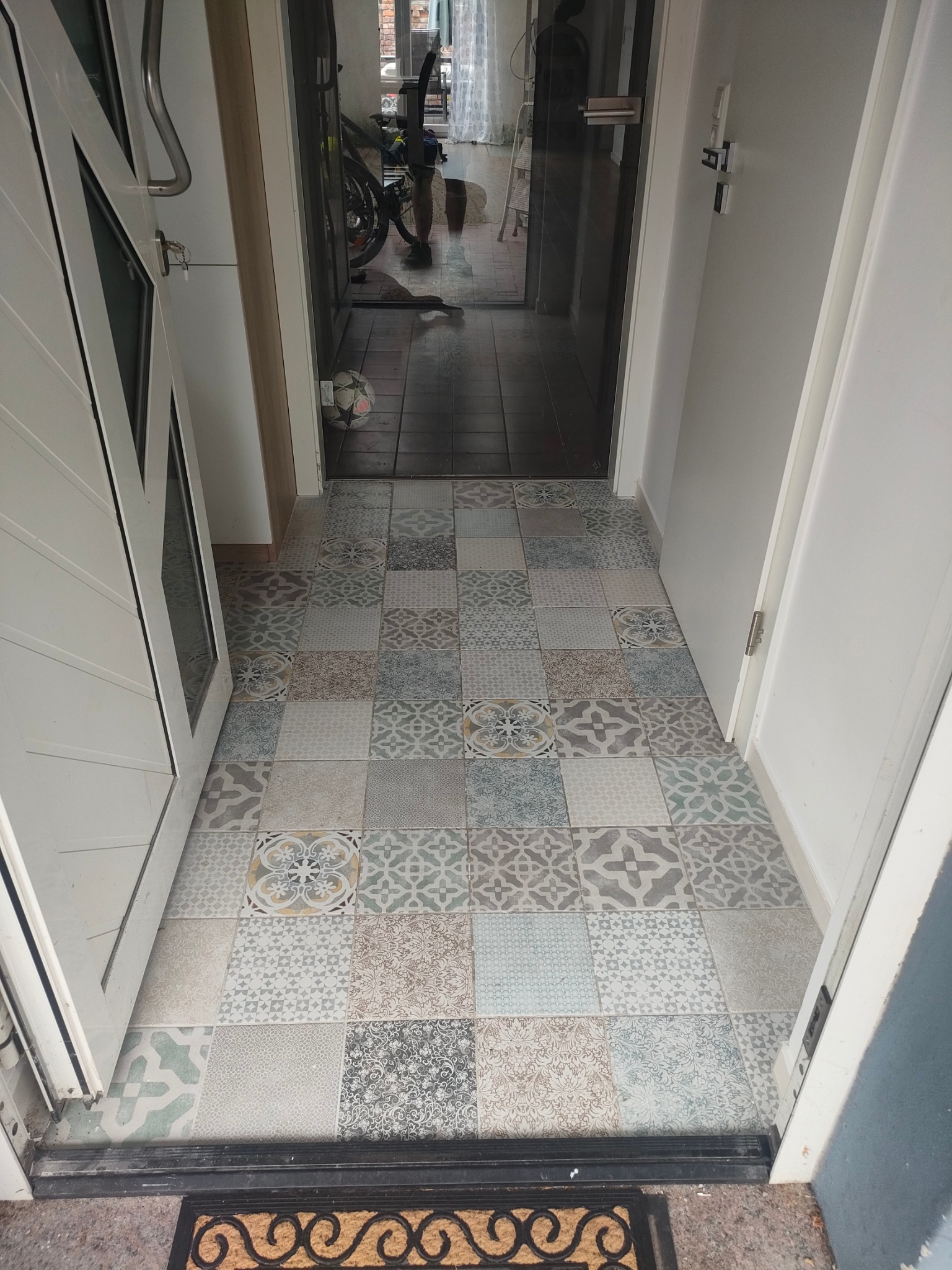 Wzorzysta, patchworkowa podłoga z płytek w przedpokoju, widok z góry. Otwarta biała rama drzwi po lewej, w tle widoczny fragment roweru i piłki.