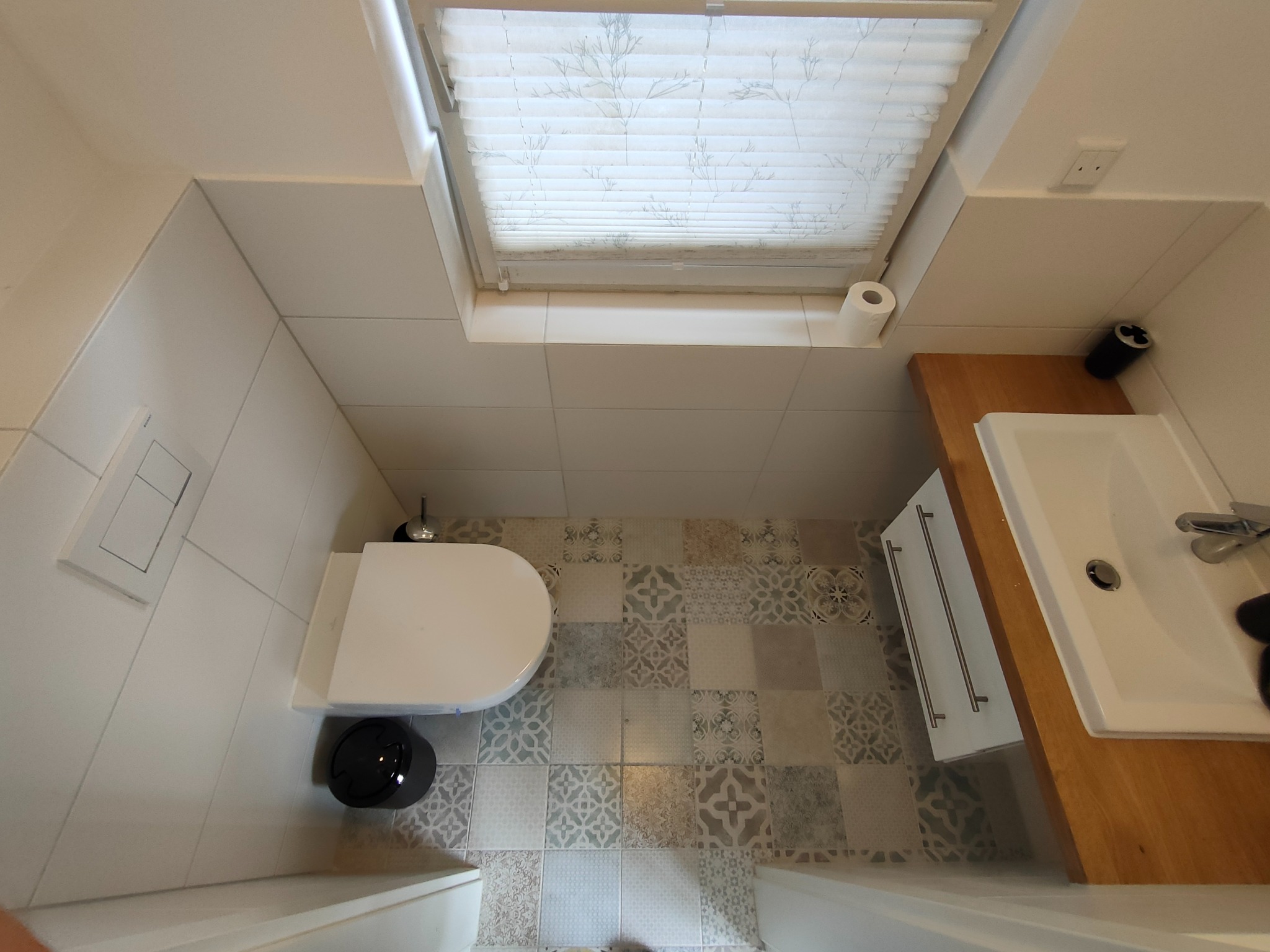 Wykończona łazienka z toaletą, umywalką z drewnianym blatem i wzorzystą podłogą z płytek. Widok z góry, okno dachowe z żaluzjami.