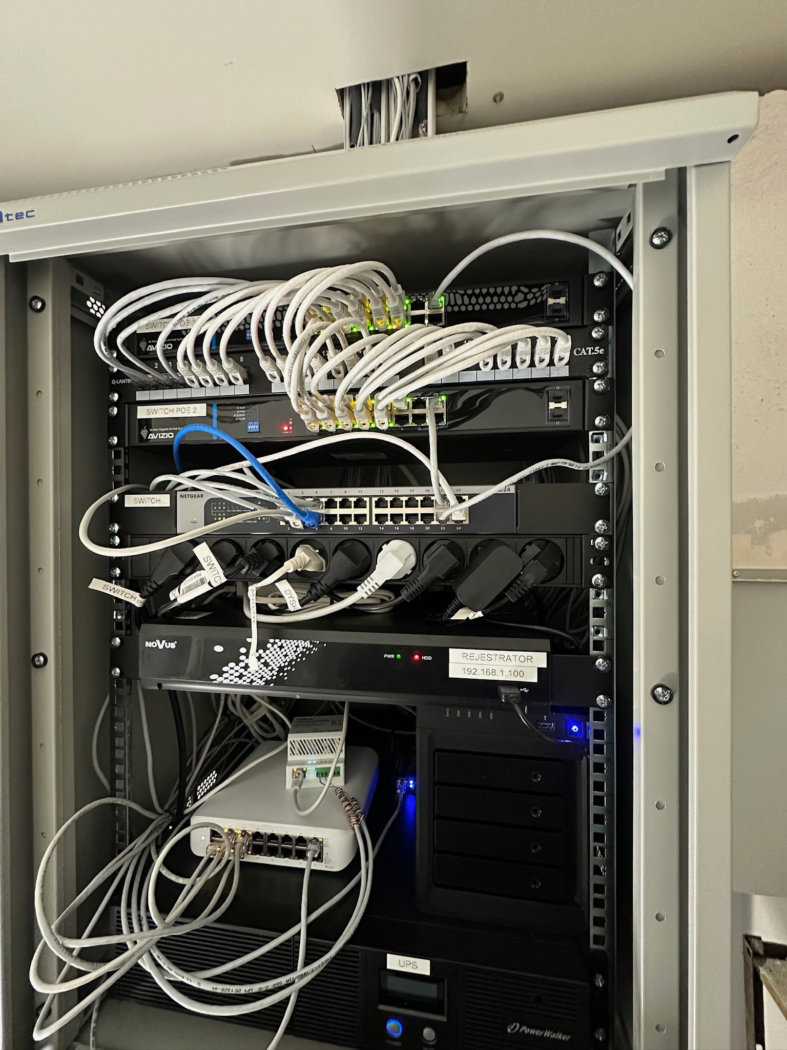 Szafa rack z zainstalowanymi urządzeniami sieciowymi: switche, rejestrator, UPS. Widoczne okablowanie, zasilacze i etykiety. Górna część szafy z otworami na kable.