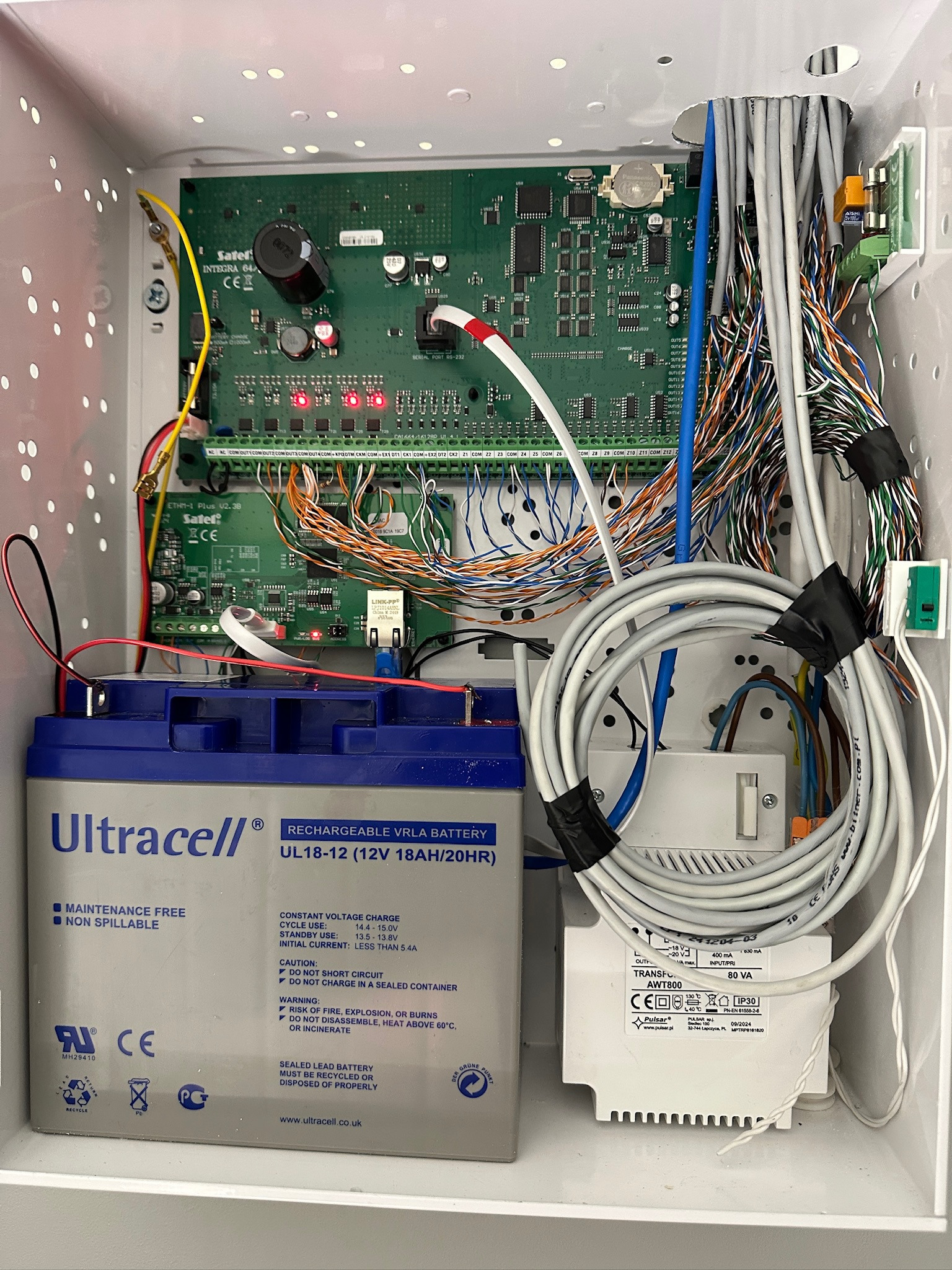 Otwarta skrzynka z elektroniką alarmową: akumulator Ultracell, transformator Pulsar AWT800, płytki Satel Integra 64, liczne przewody. Widok z góry na wnętrze instalacji.