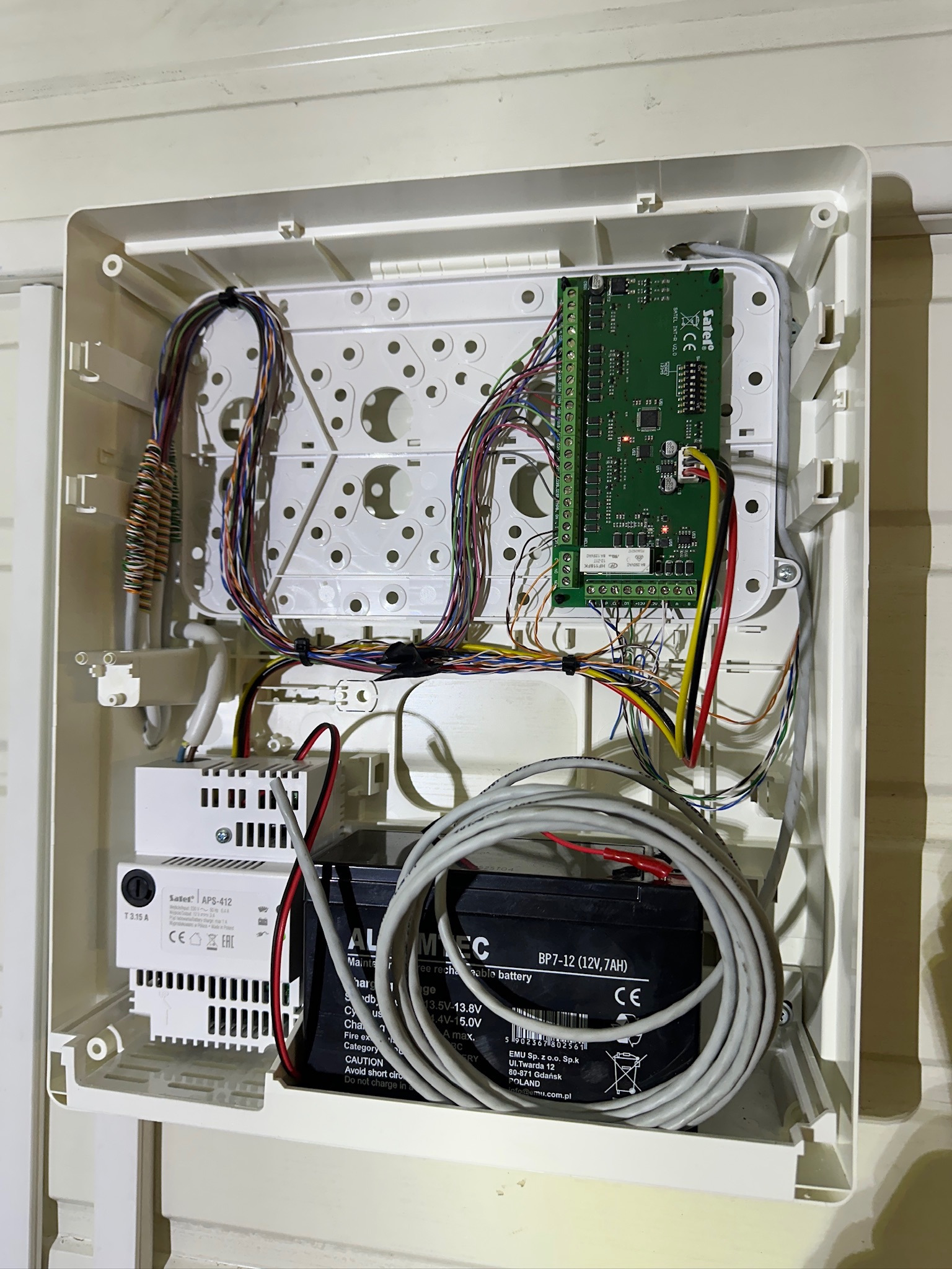 Otwarta obudowa centrali alarmowej z widoczną elektroniką, przewodami, akumulatorem i zasilaczem. Elementy systemu alarmowego wewnątrz białej skrzynki.