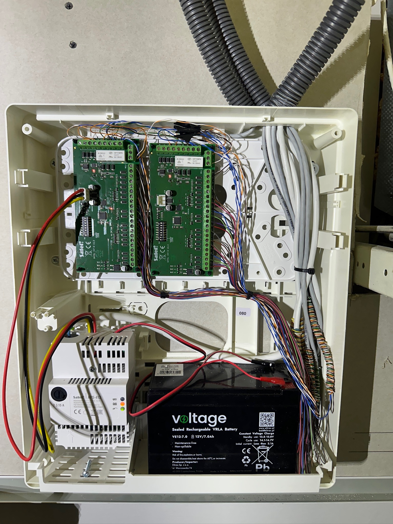 Otwarta obudowa centrali alarmowej z widocznymi modułami, przewodami i akumulatorem. Instalacja w trakcie montażu, widoczne kable i elektronika.