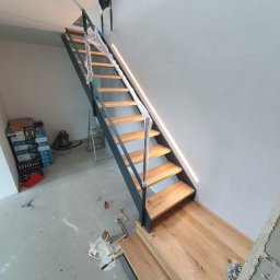 KONSTRUKCJA - Nowoczesne schody drewniane z metalową konstrukcją w surowym wnętrzu. Stopnie zabezpieczone folią, widoczne narzędzia i materiały budowlane.