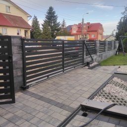 KONSTRUKCJA - Nowoczesne ogrodzenie posesji z przesuwną bramą automatyczną, wykonane z szarego betonu i poziomych, czarnych paneli metalowych. Widoczny fragment posesji i domy w tle.
