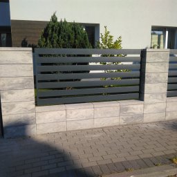 KONSTRUKCJA - Nowoczesne ogrodzenie z szarego betonu i poziomych, metalowych paneli, z bramą w tle, na tle białego domu z elementami drewna i zieleni. Widok z poziomu chodnika.
