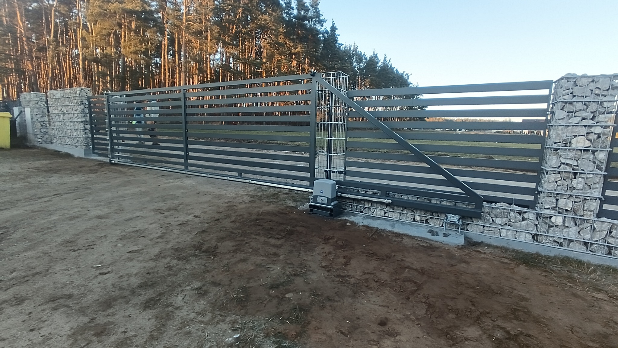 Przesuwna brama metalowa z poziomymi panelami, osadzona na słupkach z gabionów wypełnionych kamieniami. Widoczny mechanizm automatycznego otwierania bramy.