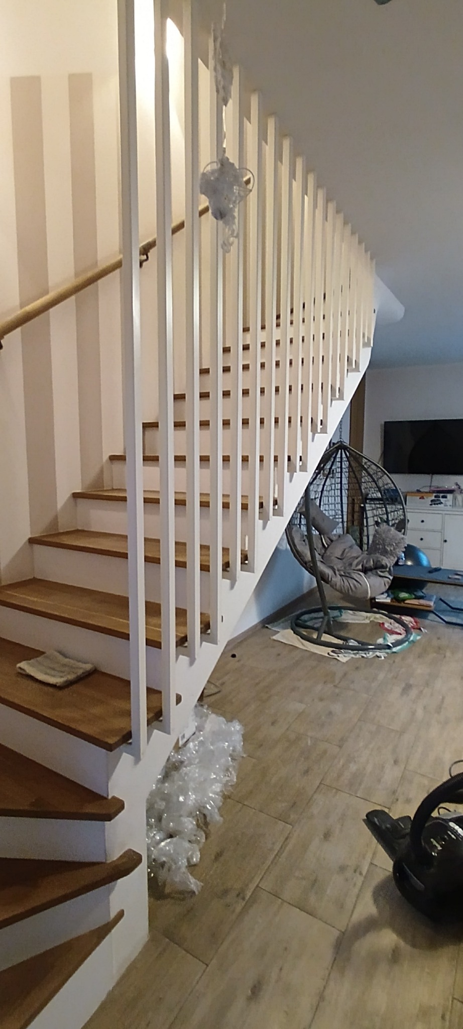 Drewniane schody z białymi barierkami w jasnym wnętrzu. Widoczne drewniane stopnie i białe pionowe elementy balustrady. W tle fotel wiszący i telewizor.