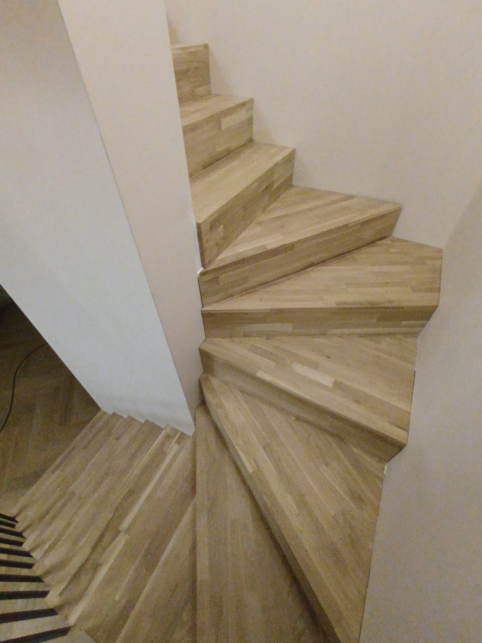 Spiralne schody drewniane z jasnego drewna, widok z góry, ukazujące unikalny kształt stopni i minimalistyczne wykończenie ściany. Detal konstrukcyjny.