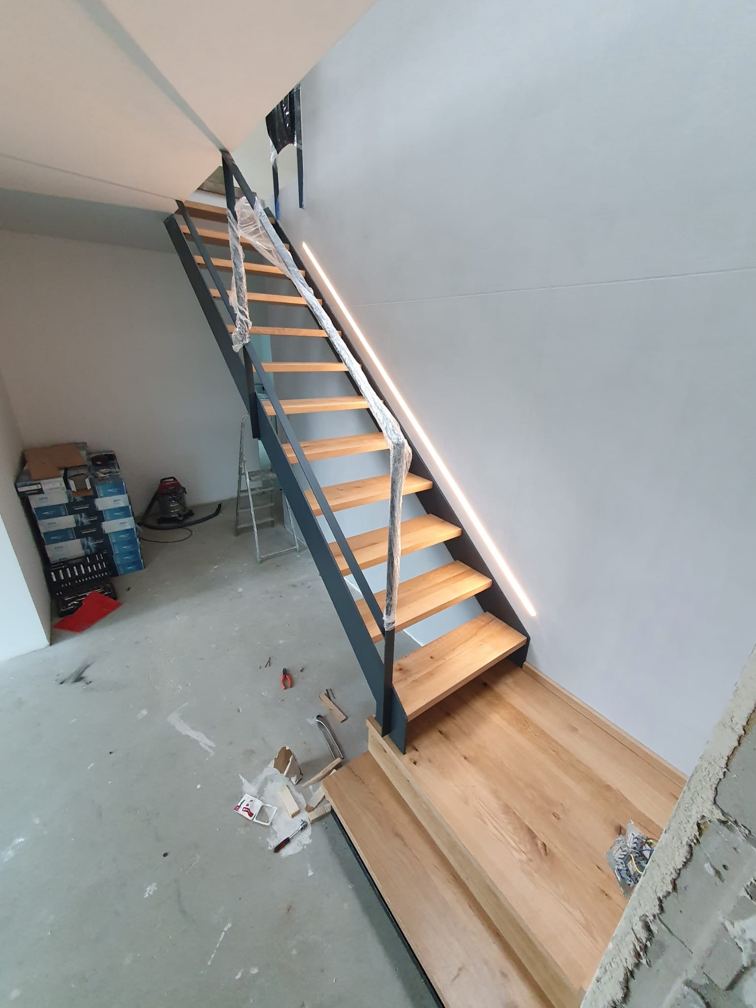 Nowoczesne schody drewniane z metalową konstrukcją w surowym wnętrzu. Stopnie zabezpieczone folią, widoczne narzędzia i materiały budowlane.