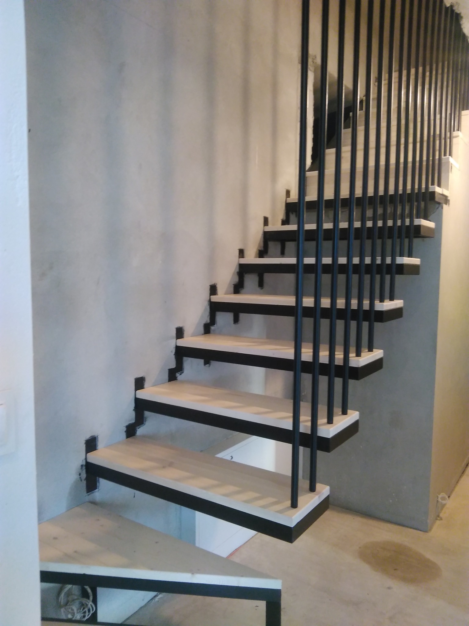 Nowoczesne schody z drewnianymi stopniami i czarną metalową konstrukcją, zamocowane do surowej, betonowej ściany. Minimalistyczny design z widocznymi elementami montażowymi.