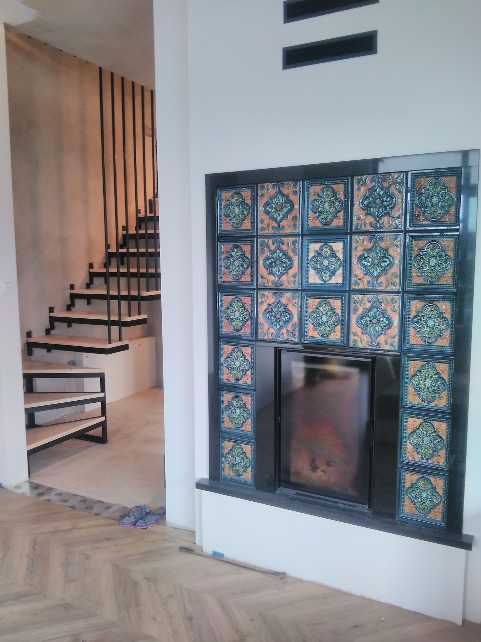 Nowoczesne schody metalowe z drewnianymi stopniami i kominek obudowany ozdobnymi kaflami w jasnym wnętrzu. Widoczny fragment parkietu ułożonego w jodełkę.