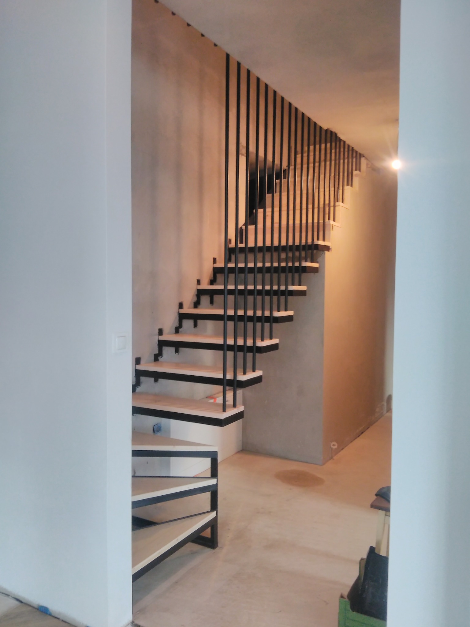 Nowoczesne schody metalowe z drewnianymi stopniami, czarne barierki pionowe, widok z dołu przez otwór w ścianie, surowy beton na ścianie, jasne oświetlenie.