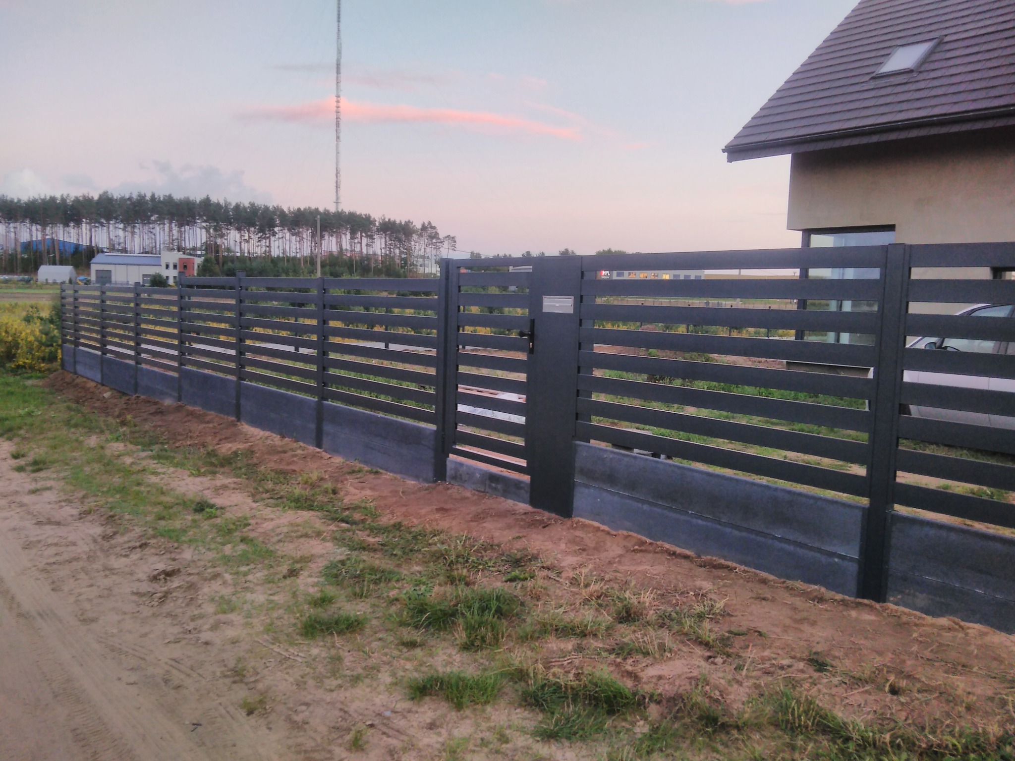 Nowoczesne, poziome ogrodzenie panelowe w kolorze antracytowym z bramką, na betonowej podmurówce. W tle dom z ciemną dachówką i drzewa.