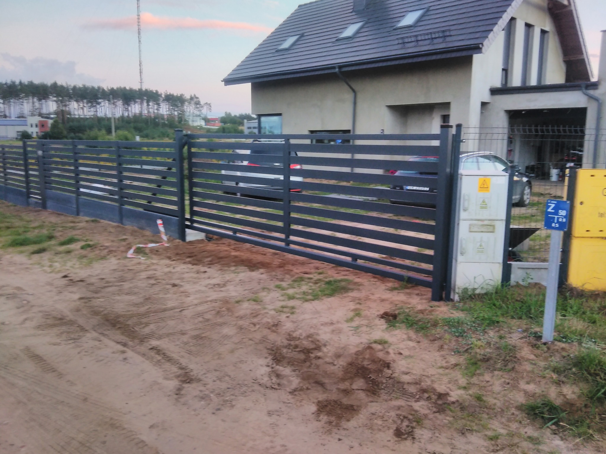 Nowoczesna, pozioma brama przesuwna z metalowymi panelami na tle domu jednorodzinnego. Widoczne skrzynki elektryczne i słupki ogrodzeniowe.