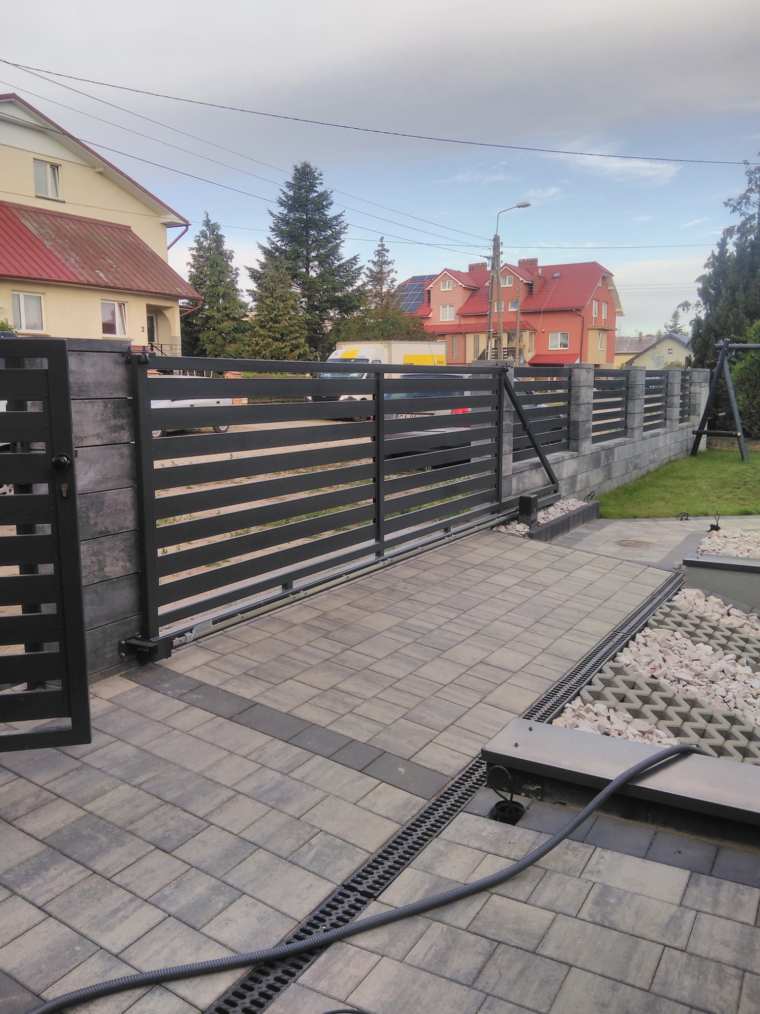 Nowoczesne ogrodzenie posesji z przesuwną bramą automatyczną, wykonane z szarego betonu i poziomych, czarnych paneli metalowych. Widoczny fragment posesji i domy w tle.