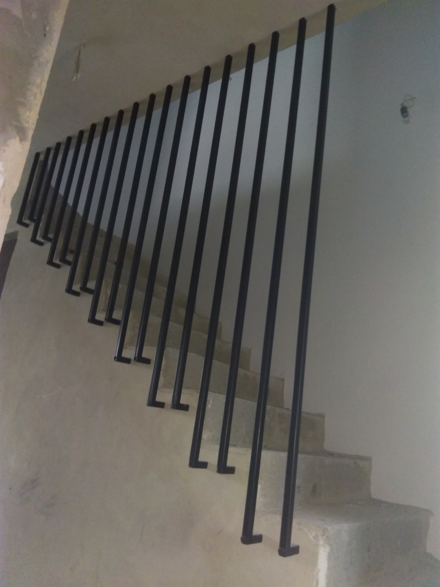 Minimalistyczne, czarne, metalowe schody z barierką w surowym wnętrzu w Rumi. Widok z dołu, podkreślający pionowe linie i prostotę konstrukcji.