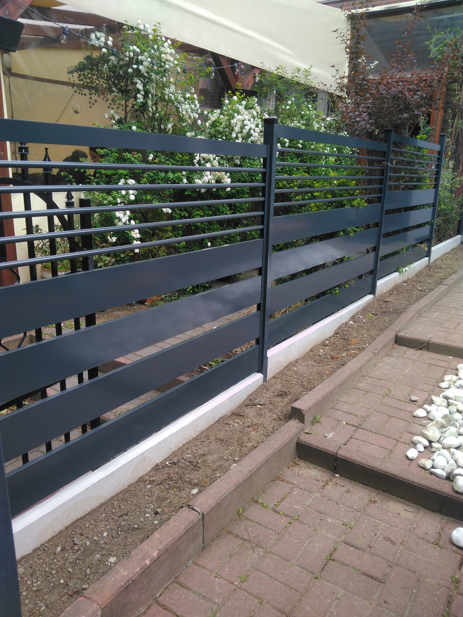 Nowoczesne, poziome ogrodzenie panelowe w kolorze grafitowym, z widoczną podmurówką i roślinnością w tle. Ułożona kostka brukowa i białe kamienie dekoracyjne.