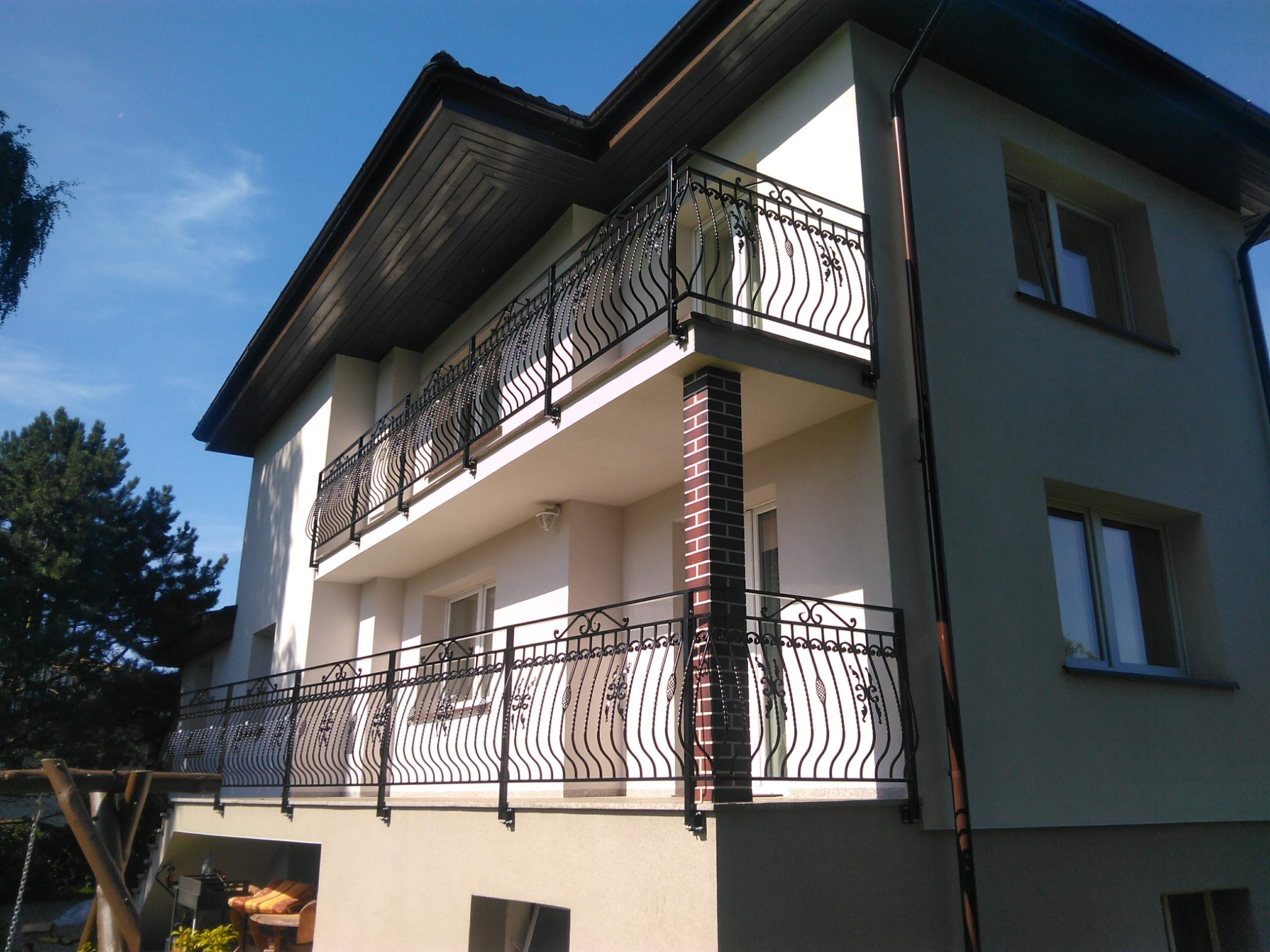Dwukondygnacyjny dom z balkonami zabezpieczonymi kutymi, czarnymi balustradami o dekoracyjnym wzorze. Widok z dołu, błękitne niebo w tle.