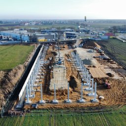 RiaBud - Widok z lotu ptaka na plac budowy z betonowymi kolumnami, dźwigiem i pracownikami w kamizelkach. W tle zabudowania miejskie i zielone pola.