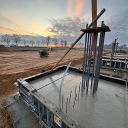 RiaBud - Zalewanie betonem szalunku z wystającymi prętami zbrojeniowymi na placu budowy o zmierzchu. W tle ubijarka i drzewa.