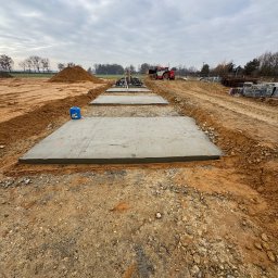 RiaBud - Wylane, betonowe płyty fundamentowe na placu budowy, widoczne z perspektywy ziemi. W tle maszyna budowlana i sterta ziemi pod pochmurnym niebem.