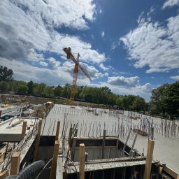 RiaBud - Zalany beton na fundamencie z wystającymi prętami zbrojeniowymi, widok z góry na plac budowy z żurawiem i błękitnym niebem w tle.