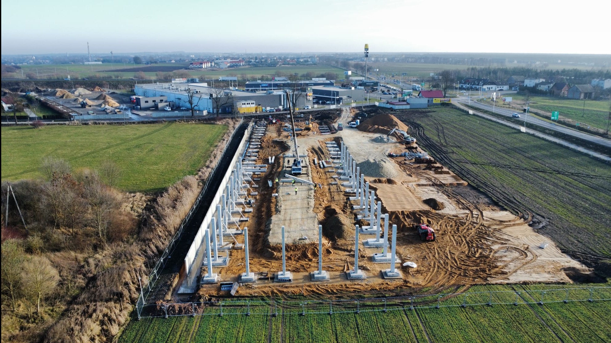 Widok z lotu ptaka na plac budowy z betonowymi kolumnami, dźwigiem i pracownikami w kamizelkach. W tle zabudowania miejskie i zielone pola.