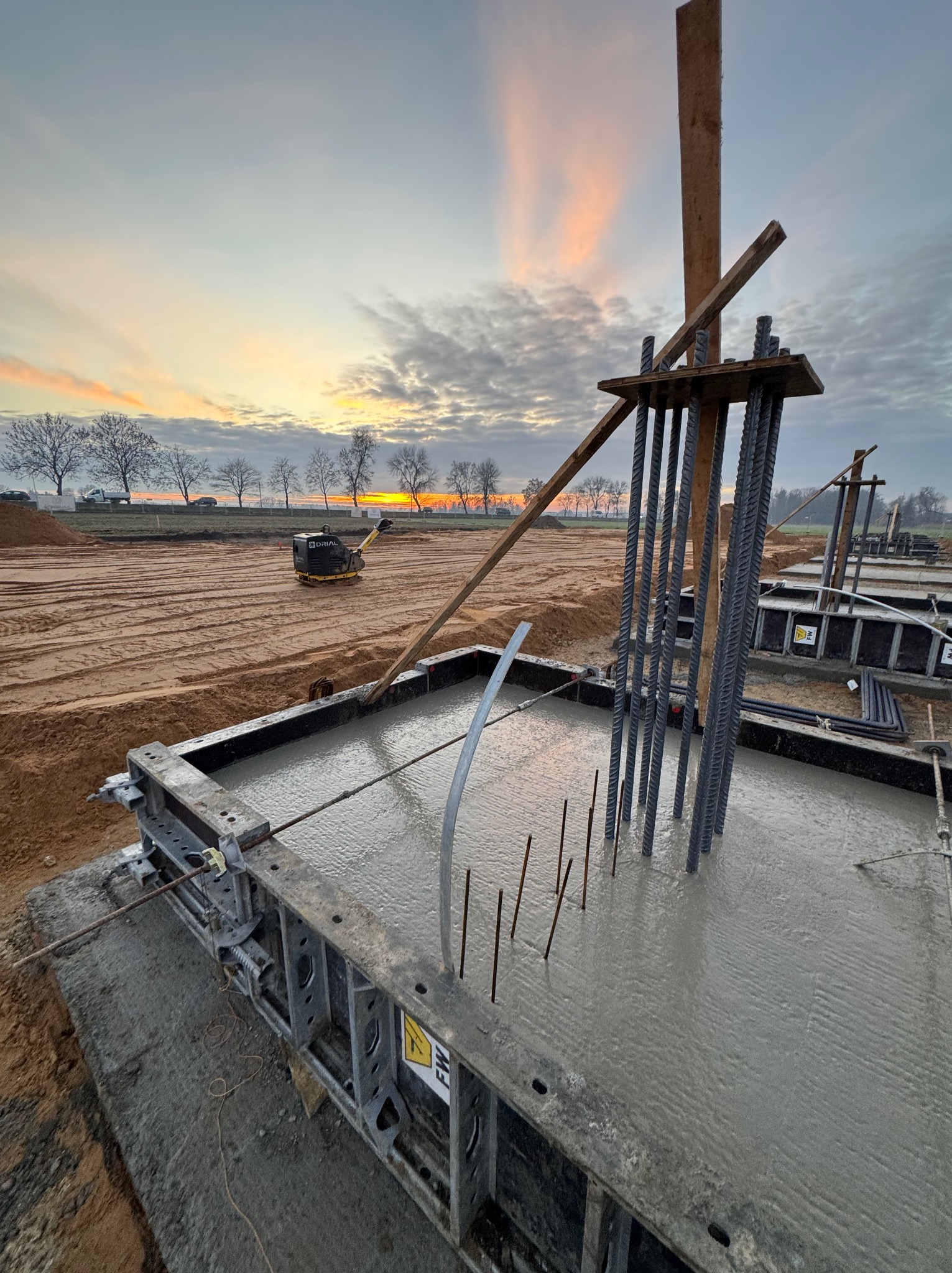 Zalewanie betonem szalunku z wystającymi prętami zbrojeniowymi na placu budowy o zmierzchu. W tle ubijarka i drzewa.