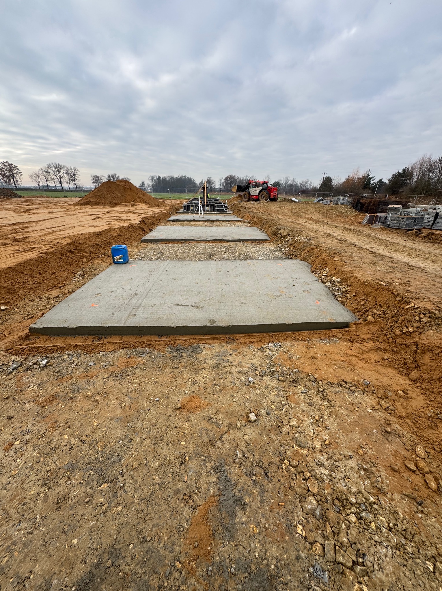 Wylane, betonowe płyty fundamentowe na placu budowy, widoczne z perspektywy ziemi. W tle maszyna budowlana i sterta ziemi pod pochmurnym niebem.