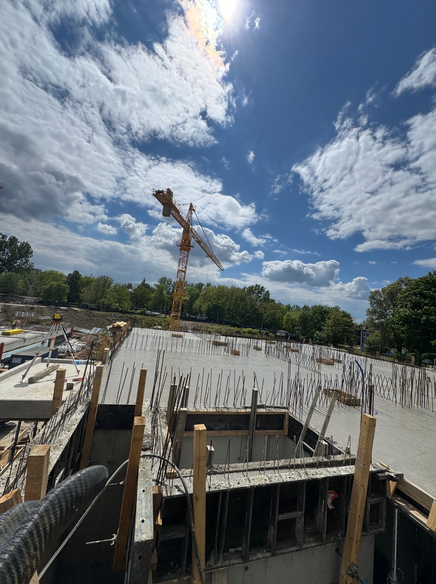 Zalany beton na fundamencie z wystającymi prętami zbrojeniowymi, widok z góry na plac budowy z żurawiem i błękitnym niebem w tle.