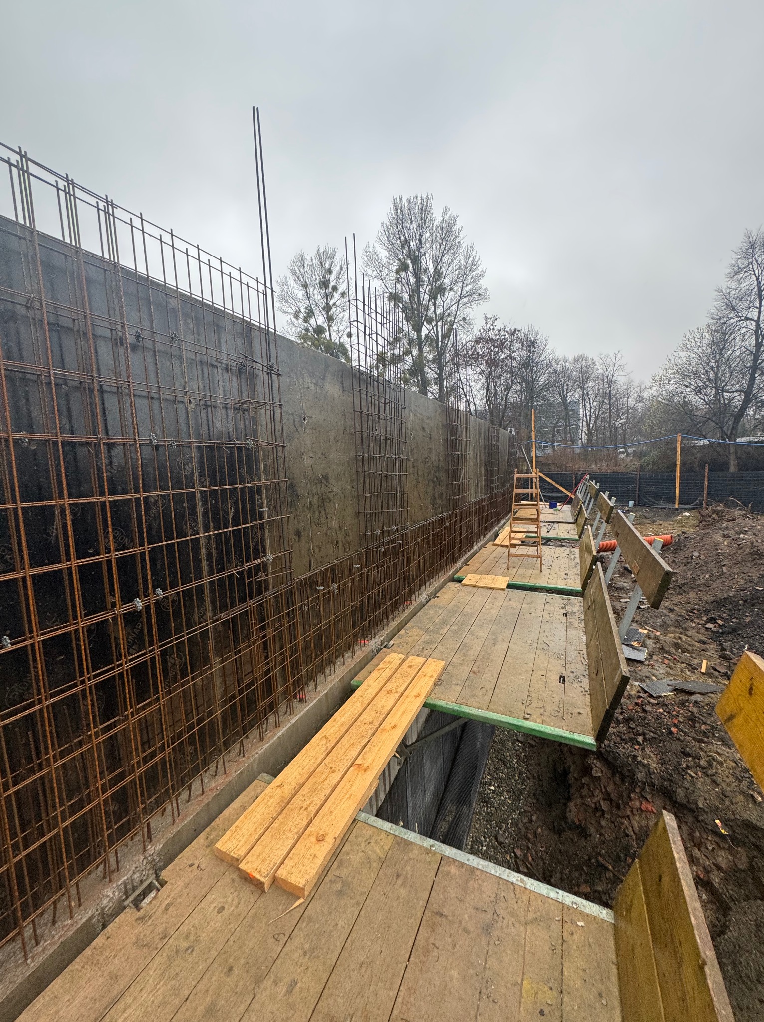 Zbrojenie pionowe fundamentów z deskowaniem i pomostem roboczym. Widoczne stalowe pręty zbrojeniowe, szalunki i drewniane deski pomostu. Pochmurne niebo w tle.