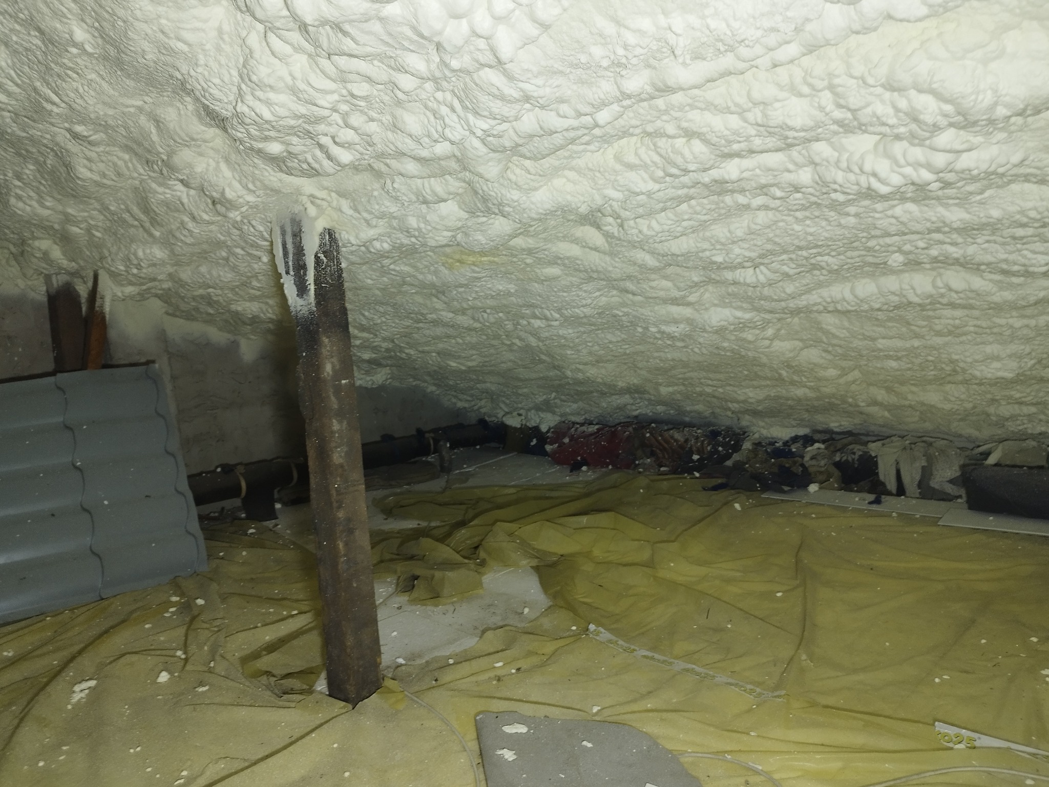 Poddasze po natrysku pianki PUR, widoczne drewniane elementy konstrukcyjne i folia ochronna na podłodze. Biała, nierówna powierzchnia izolacji.