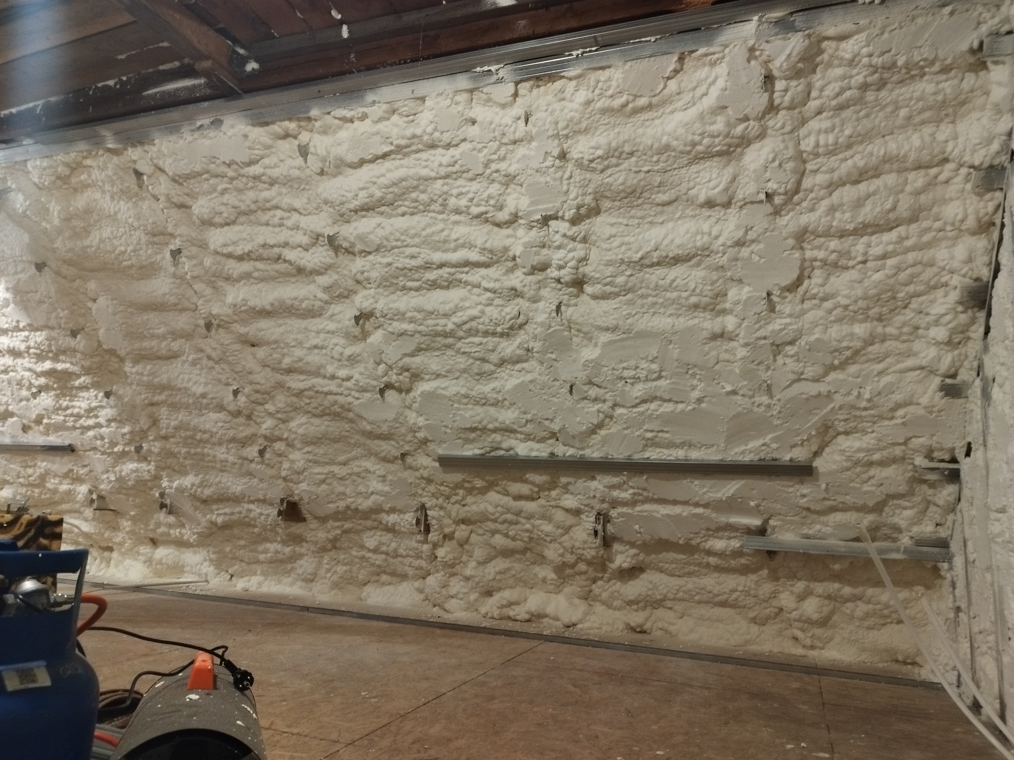 Ściana pokryta grubą warstwą białej piany izolacyjnej, z widocznymi metalowymi elementami konstrukcyjnymi i sprzętem do natrysku w dolnej części kadru.