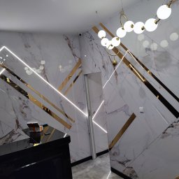 Kamil Skibicki - Eleganckie wykończenie wnętrza z marmurowymi płytami, złotymi akcentami i nowoczesnym oświetleniem. Widoczny fragment czarnego mebla i drzwi. Nietypowy układ płyt.