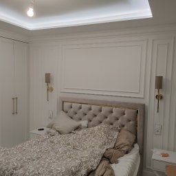 Kamil Skibicki - Elegancka sypialnia w bieli: łóżko z tapicerowanym zagłówkiem, ozdobne listwy ścienne, symetryczne kinkiety i lustrzana szafa. Subtelne oświetlenie sufitowe.