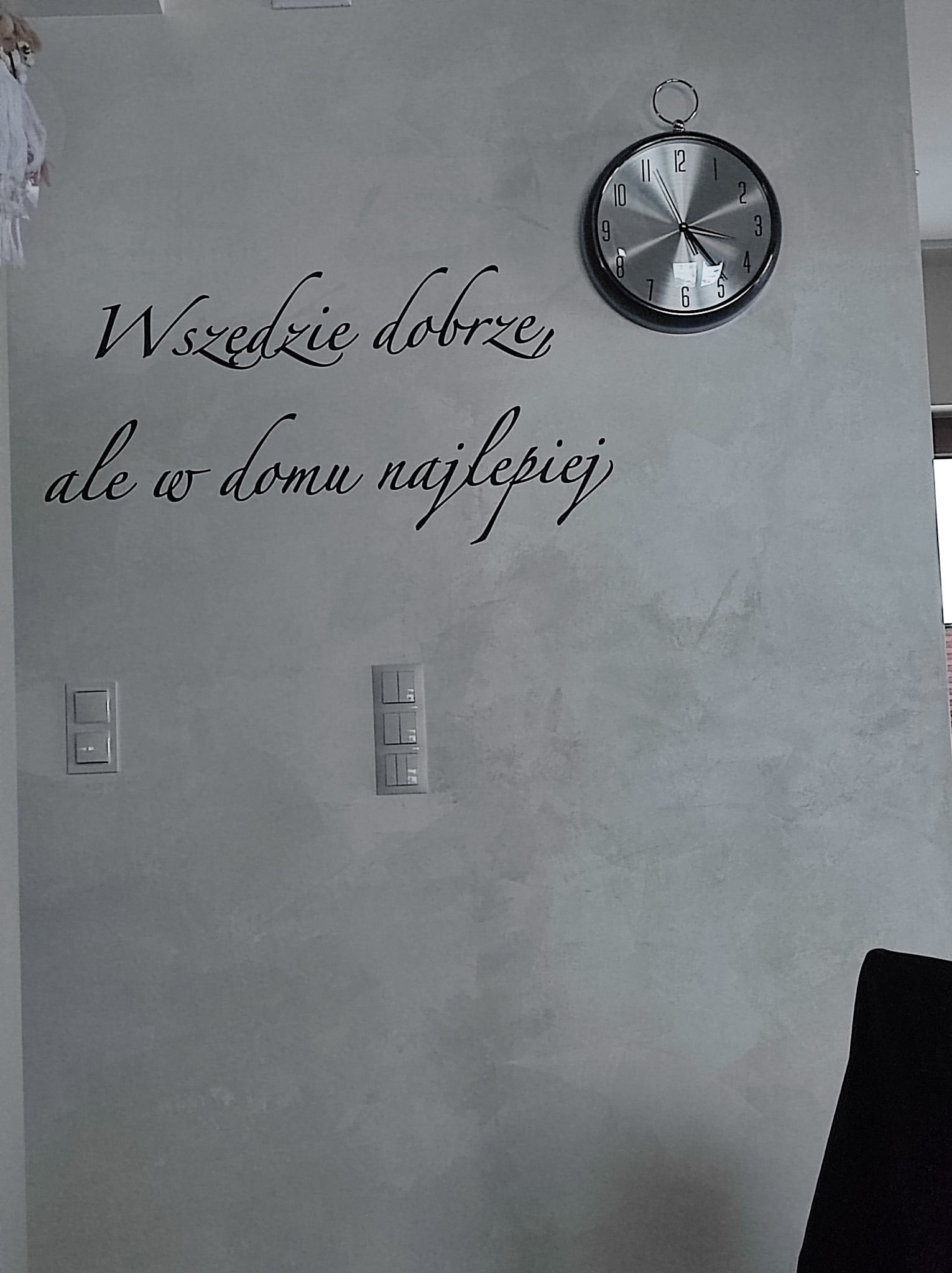 Ściana w salonie po szpachlowaniu z dekoracyjnym napisem i zegarem. Widoczne włączniki światła. Delikatna faktura ściany.