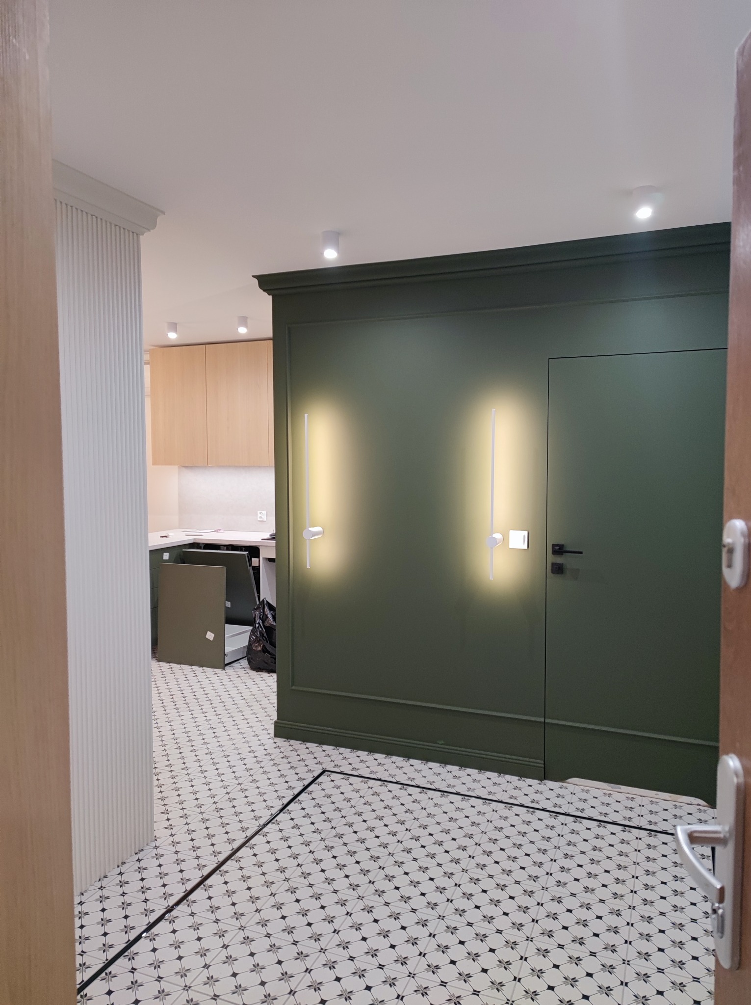 Nowoczesne wnętrze: oliwkowa ściana z listwami, geometryczna podłoga, minimalistyczne oświetlenie. Widok zza drzwi, częściowo widoczna kuchnia w tle.