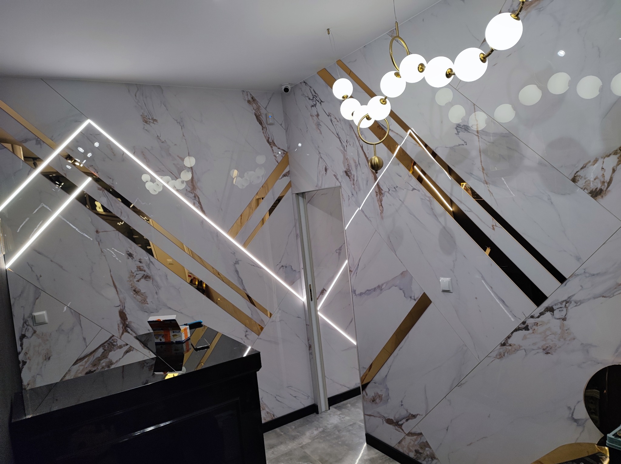 Eleganckie wykończenie wnętrza z marmurowymi płytami, złotymi akcentami i nowoczesnym oświetleniem. Widoczny fragment czarnego mebla i drzwi. Nietypowy układ płyt.
