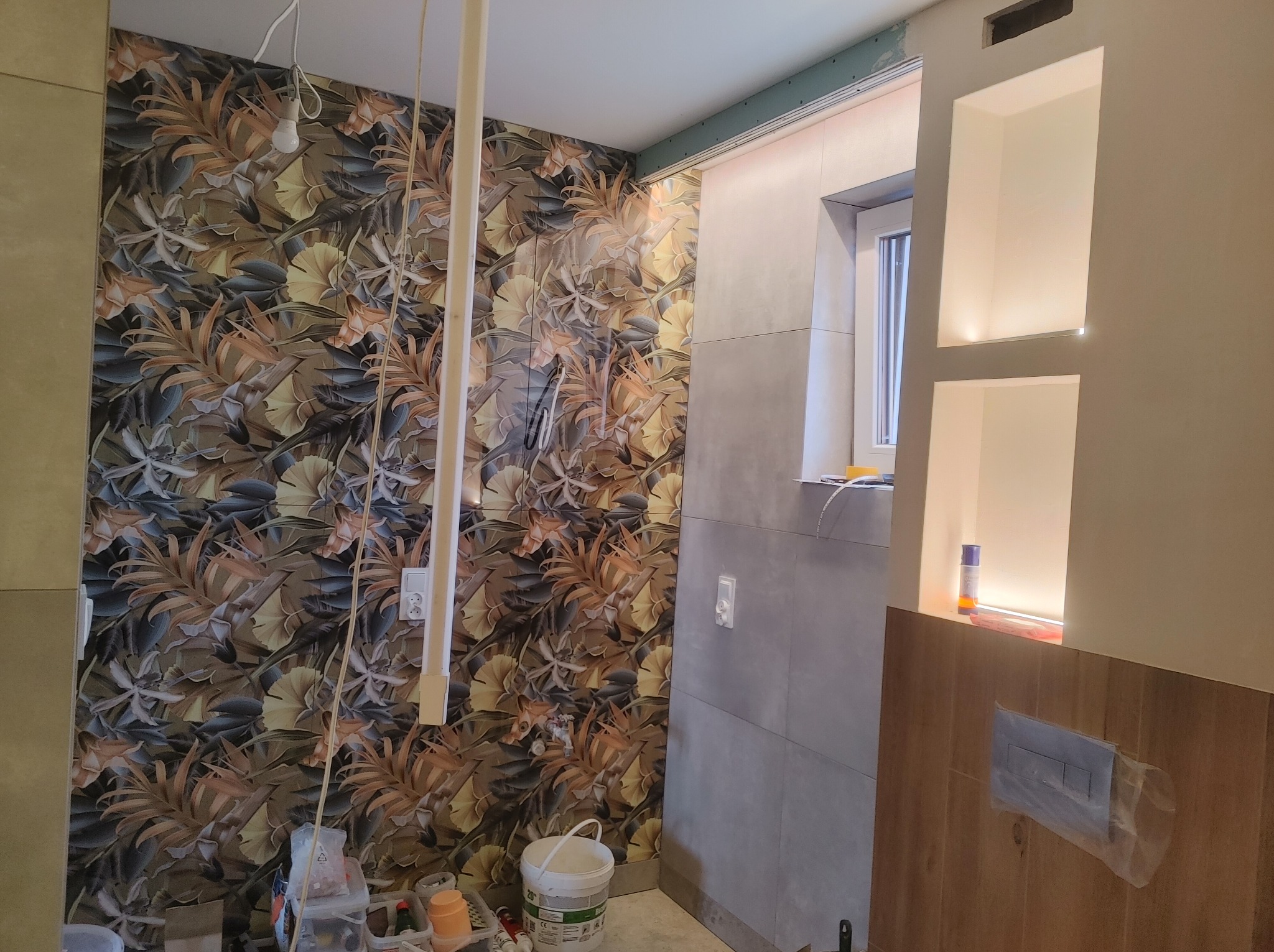 Wykończenie łazienki w trakcie: ściana z wzorzystą tapetą, płytki imitujące beton, wnęki z oświetleniem LED. Widoczne narzędzia i materiały budowlane.