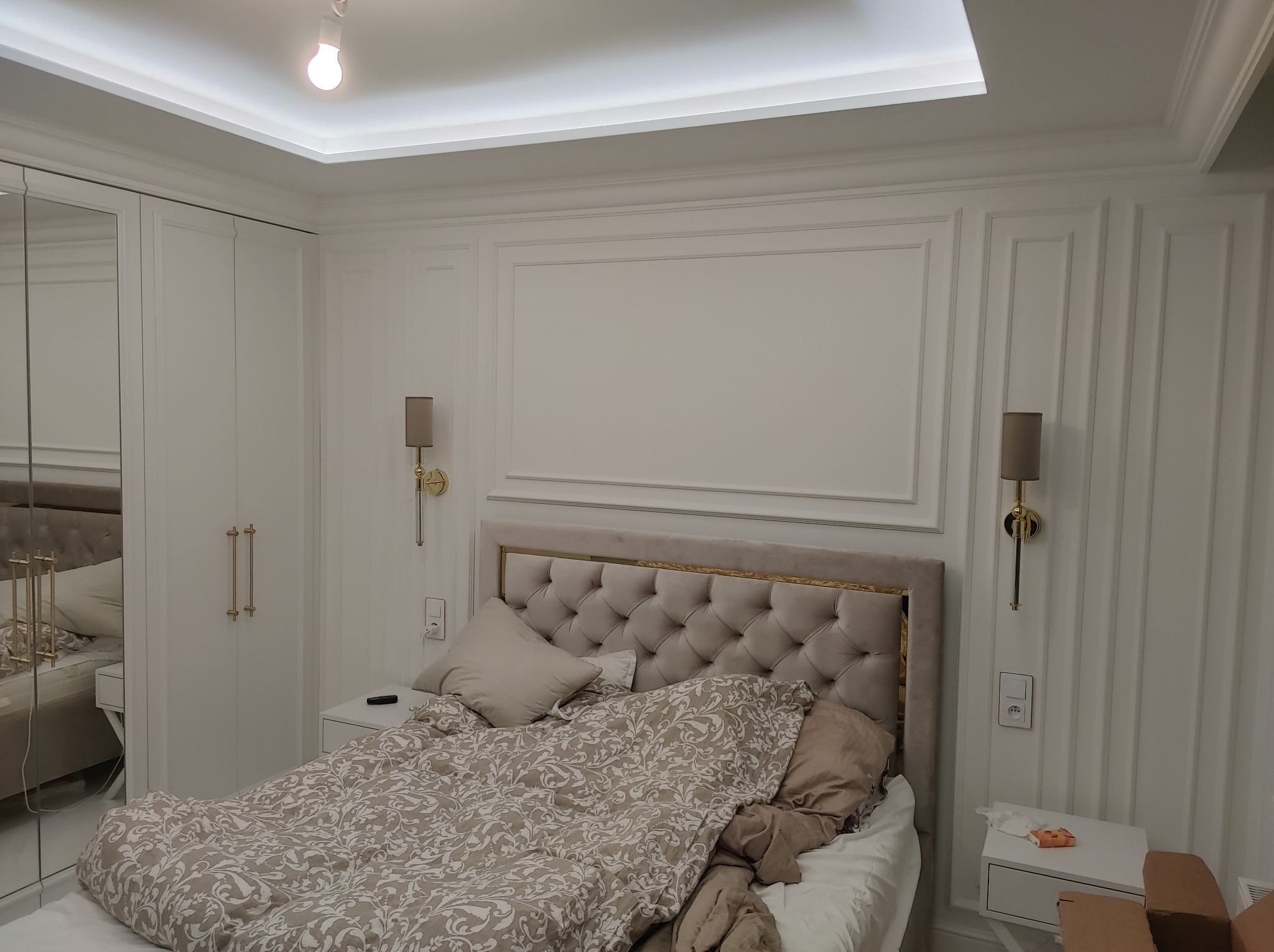 Elegancka sypialnia w bieli: łóżko z tapicerowanym zagłówkiem, ozdobne listwy ścienne, symetryczne kinkiety i lustrzana szafa. Subtelne oświetlenie sufitowe.