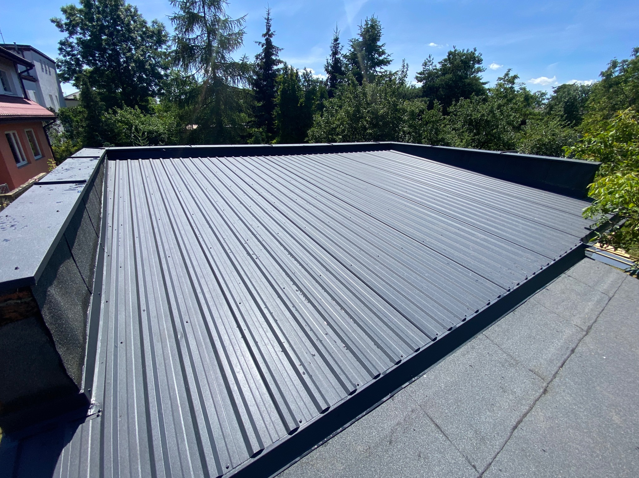 Szary, blaszany dach z widocznymi łączeniami, na tle zieleni drzew i fragmentu budynku. Widok z góry, w słoneczny dzień.